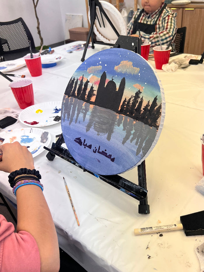 Person painting a scenic round canvas on a table with art supplies.